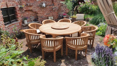 A Teak 180cm Maximus Round Set (8 San Francisco Chairs) featuring a round table with chairs, situated in a brick courtyard garden, surrounded by lush plants and flowers.