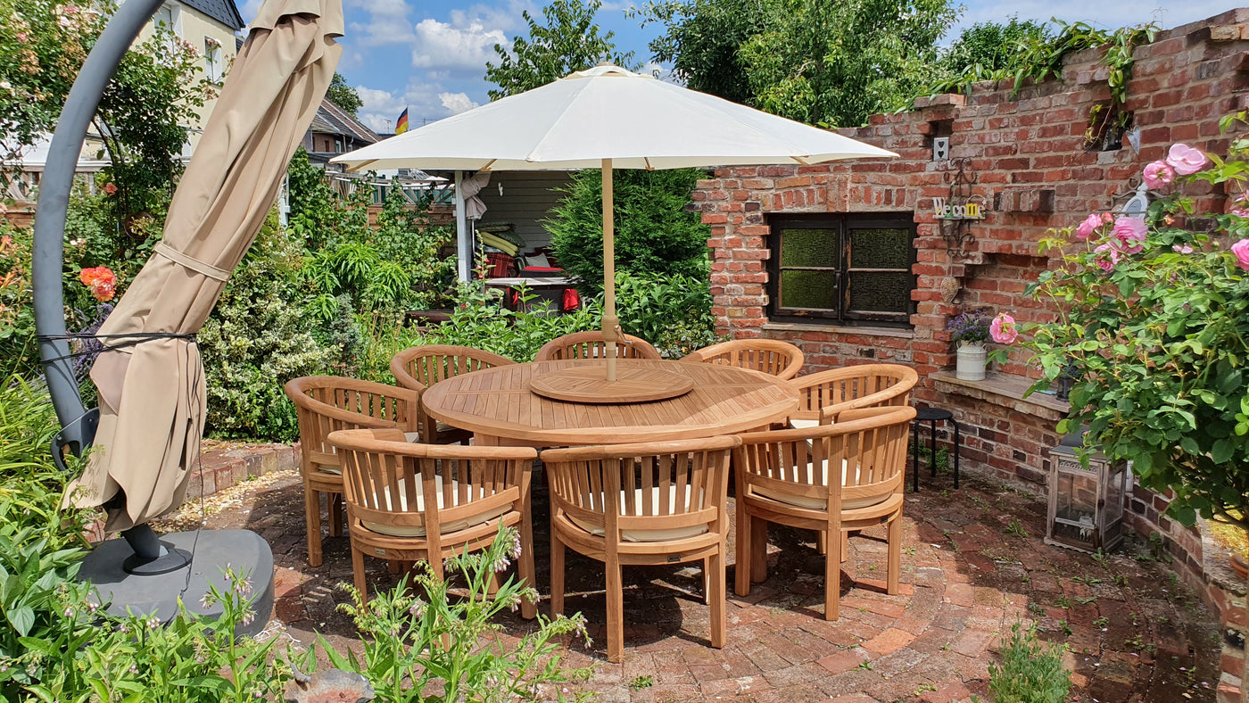 Teak 180cm Maximus Round Set (8 San Francisco Chairs) Complete set Cushions included with chairs under a white umbrella, set in a garden with brick walls and blooming roses, sunny day.