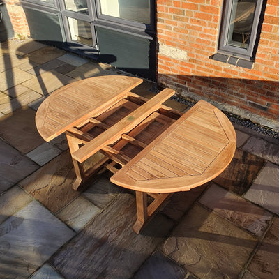 A Teak Garden Furniture Round To Oval 120-170cm Extending Table with a split top, partially folded, on a stone-paved patio near a brick building.