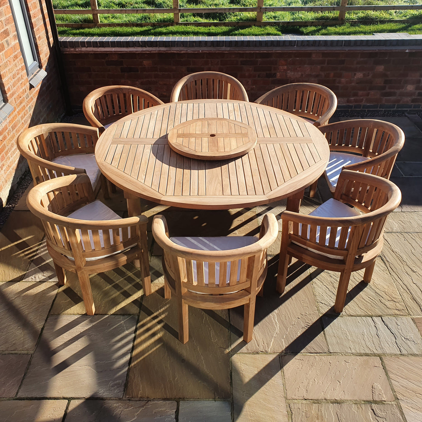 Round Teak 180cm Maximus Patio Dining Table Set (8 San Francisco Chairs) Complete set Cushions included, set on a stone terrace under sunlight, surrounded by a brick wall.