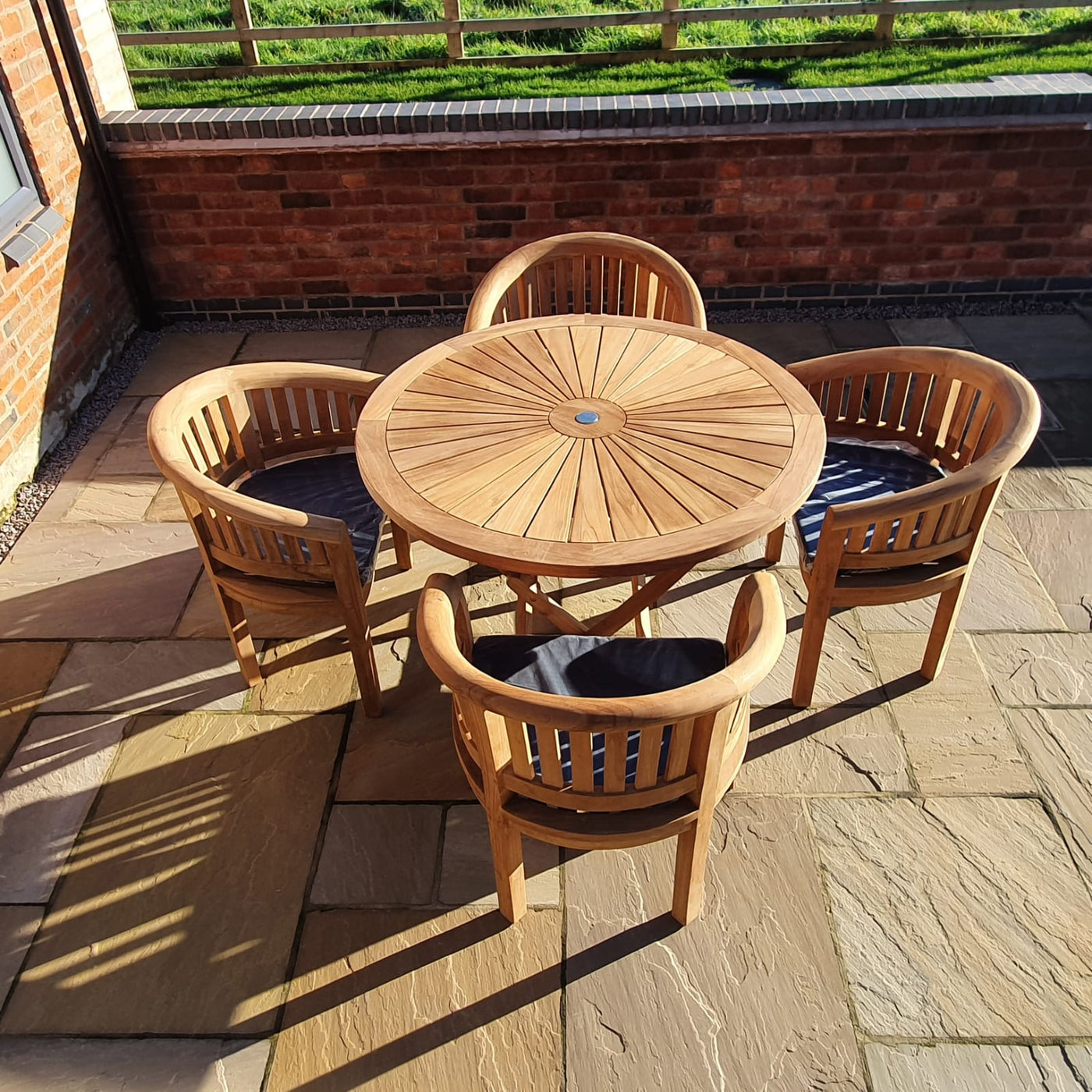 Teak 120cm Sunshine Set with 4 San Francisco Chairs includes a round table and four chairs on a paved patio, sunlight casting shadows, surrounded by a brick wall.