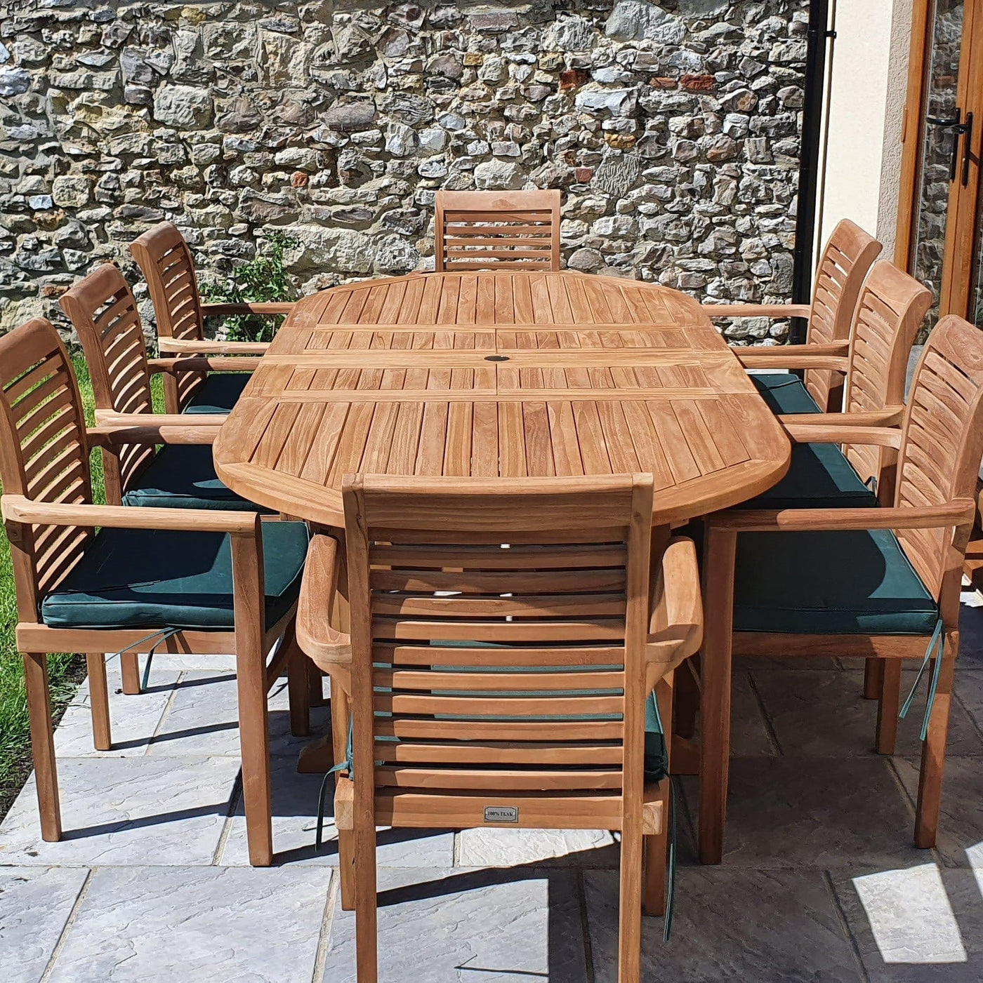 A Premium Teak Oval 180-240cm Garden Furniture Set with 8 Oxford Stacking Chairs, cushions included, set on a patio against a stone wall.