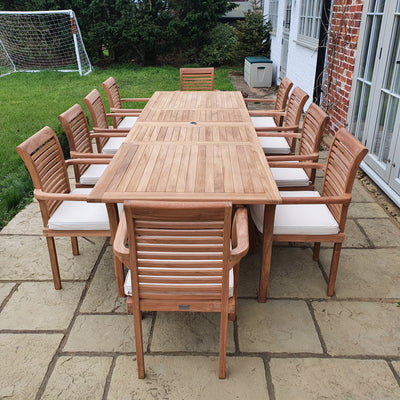 Teak 2-3m Extending Rectangle Table (10 Seat Oxford Stacking Set) complete set with cushions included, on a stone patio adjacent to a white house and green lawn.