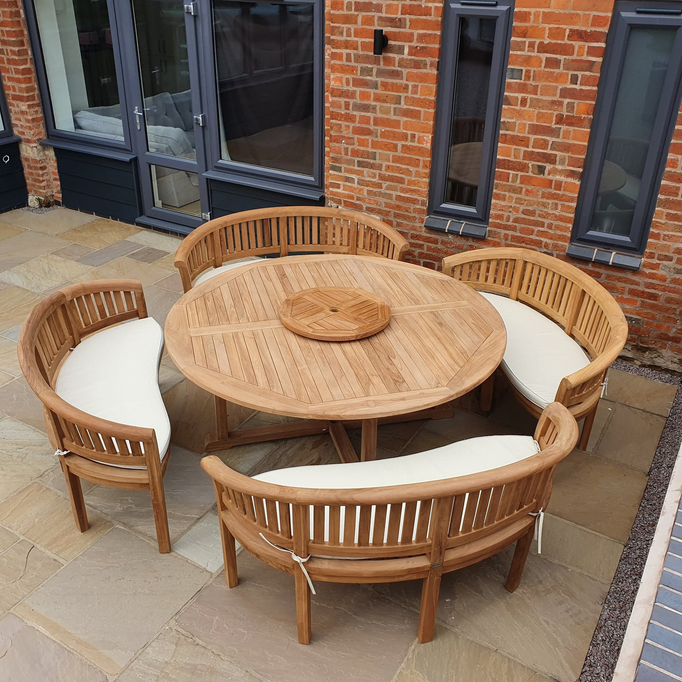 A Teak 180cm Round Maximus Set (4 San Francisco Benches) complete set with cushions on a brick patio, with a backdrop of a brick building and glass doors.