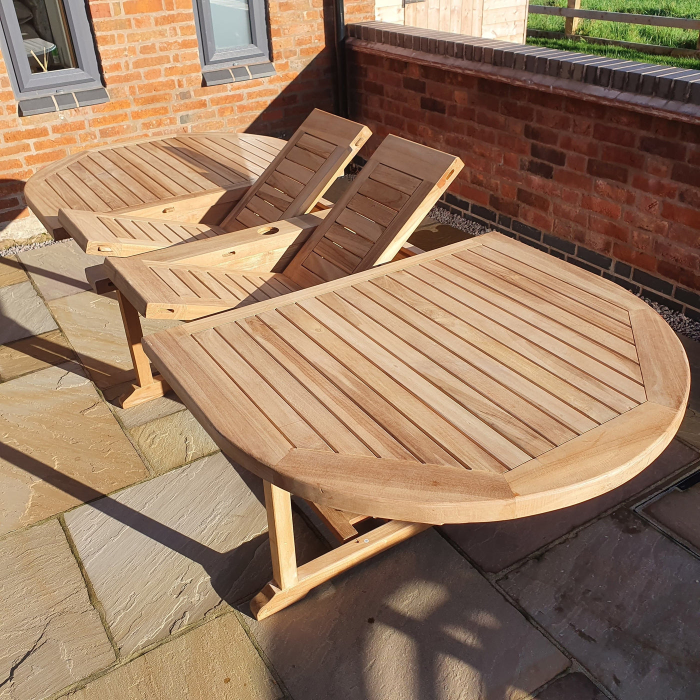 Two Premium Teak Oval 180-240cm garden furniture sets (with 8 Oxford stacking chairs) cushions included on a brick patio, bathed in sunlight.