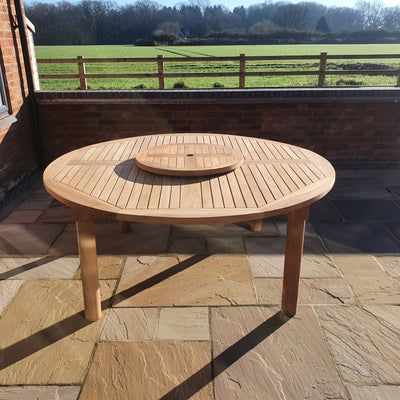 A Teak 180cm Maximus Round Stacking Set with 8 Oxford Stacking Chairs on a stone patio, overlooking a rural landscape with a clear sky and fenced fields.