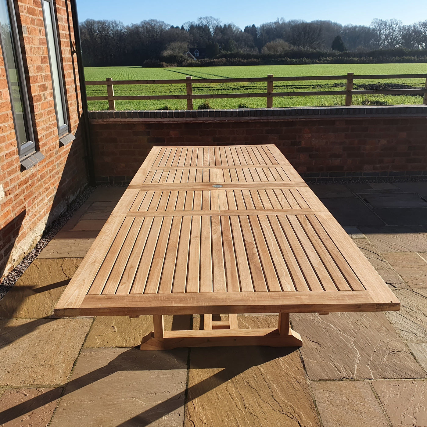 A Giant 2-3m Teak Extending Rectangle Table with 12 Oxford Stacking Chairs, complete set and cushions included, on a brick patio beside a red brick building, with a grassy field and trees in the background on a sunny day.