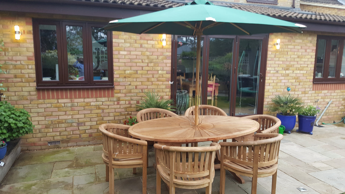 Teak 2m sunshine with 6 San Francisco Chairs & cushions garden furniture set with a round table and chairs under a green umbrella on a brick terrace, adjacent to a house with large windows.