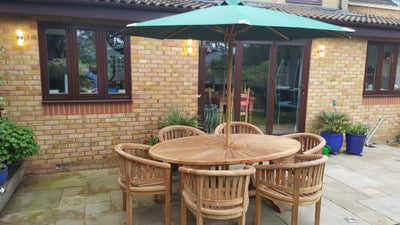 Teak 2m sunshine with 6 San Francisco Chairs & cushions garden furniture set with a round table and chairs under a green umbrella on a brick terrace, adjacent to a house with large windows.