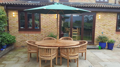 Outdoor dining area with a Teak 2m sunshine table and 6 San Francisco Chairs & cushions under a green umbrella, located on a brick patio against a brick house with large windows.