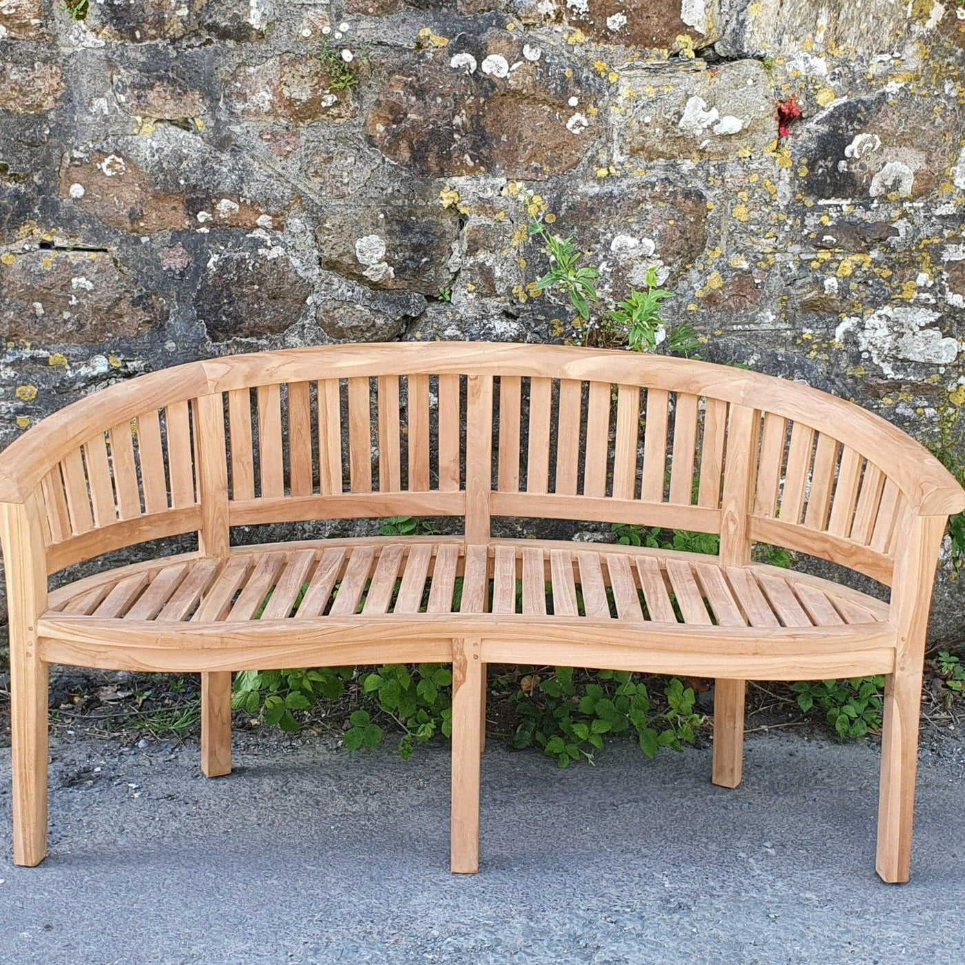 Teak 180cm Round Maximus Set (3 San Francisco Benches) Complete set Cushions Included curved bench in front of a weathered stone wall with small plants growing at its base.