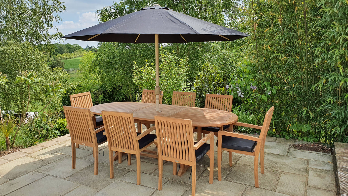 Teak 180-240cm Oval Extending Table with 8 Henley Stacking Chairs set under a large umbrella, surrounded by lush greenery and overlooking a countryside landscape.