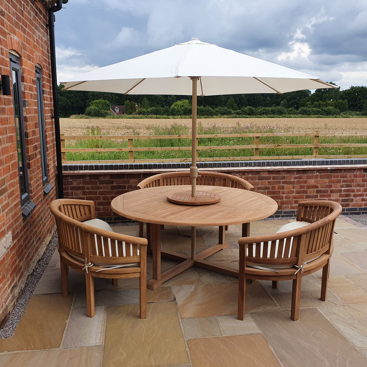 Teak 180cm Round Maximus Set (3 San Francisco Benches) Complete set Cushions Included table with chairs under a large white umbrella on a patio beside a brick building, overlooking a field.