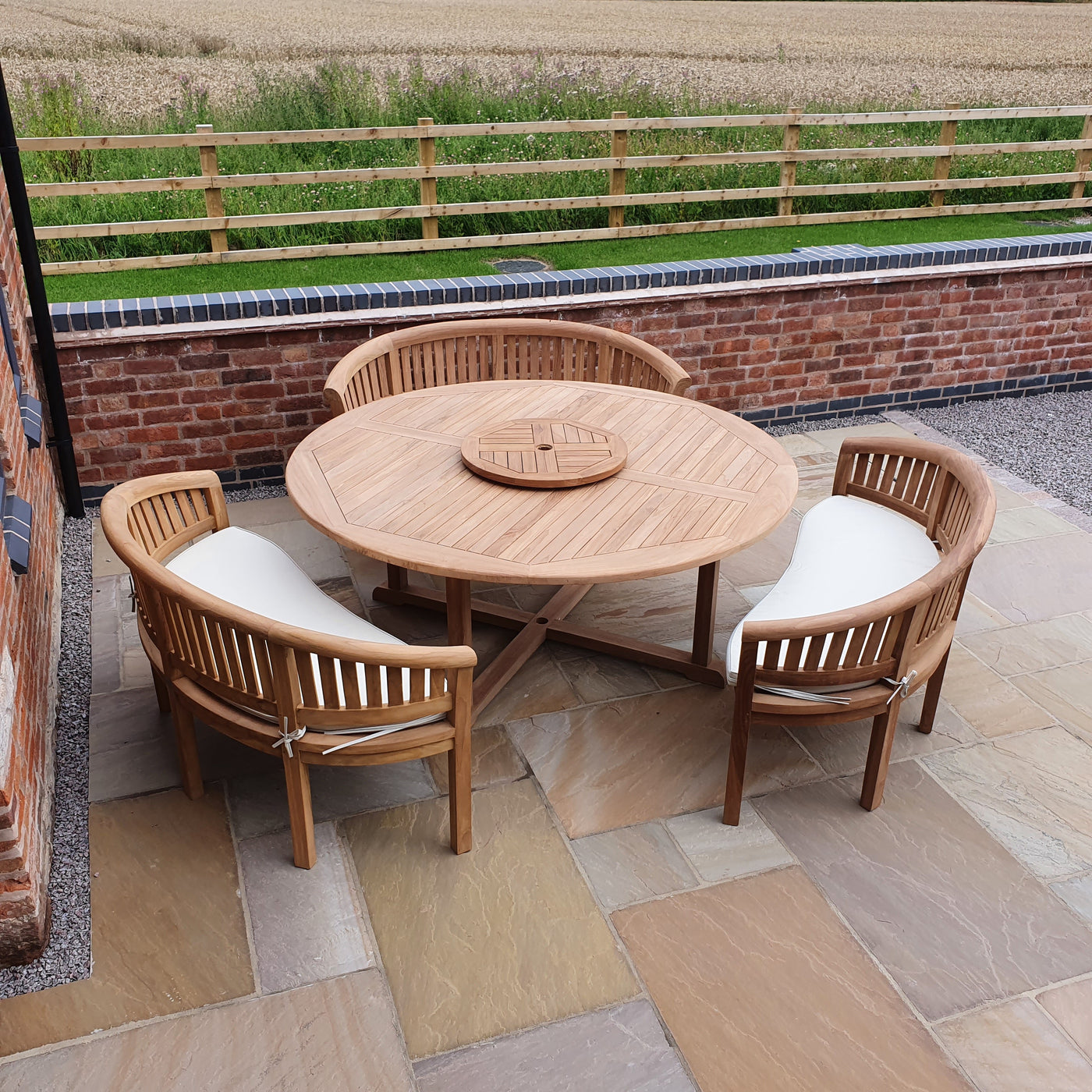 Outdoor dining setup featuring a Teak 180cm Round Maximus Set with 3 San Francisco Benches and cushions on a stone patio, overlooking a countryside field.