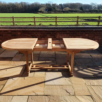 A Teak 180-240cm Oval Extending Table with 8 Henley Stacking Chairs on a stone patio, overlooking a grassy field with trees and a clear sky.