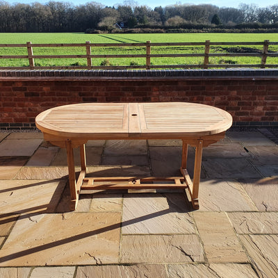 A Teak 180-240cm Oval Extending Table with 8 Henley Stacking Chairs on a paved patio with a countryside view and clear sky in the background.