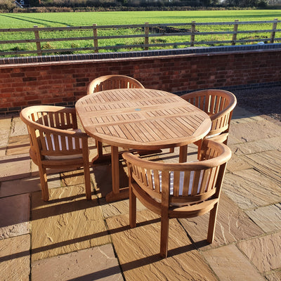 Teak Round To Oval 120-170cm Extending Table & 4 San Francisco Chairs Complete set Cushions Free Delivery consisting of one round table and four chairs on a brick terrace, with a grassy field and clear blue sky in the background.