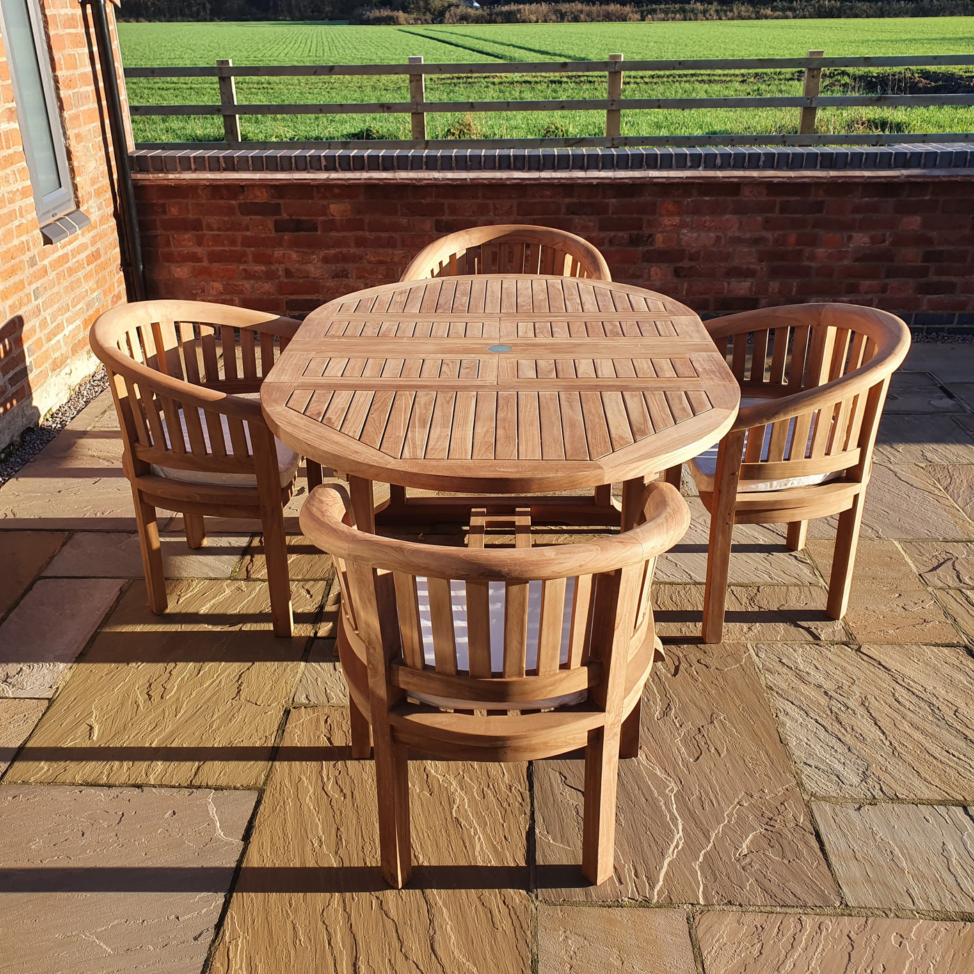 A Round To Oval 120-170cm Extending Table & 4 San Francisco Chairs Complete set Cushions Free Delivery with four matching chairs on a stone patio, sunlight casting shadows, with a grassy field and a fence in the background.