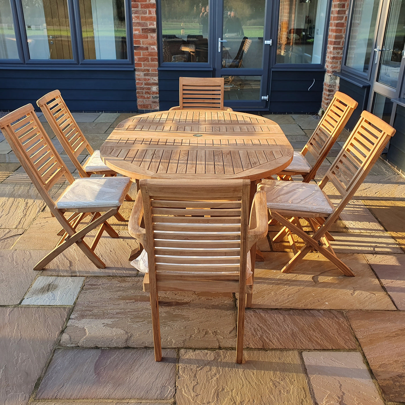 Oval 120-170cm Extending 2 Seat Stacking & 4 Folding Chair Complete set Cushions Free Delivery round table and four chairs on a paved patio, sunny day, near a building with brick walls and large windows.