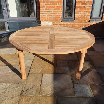 Round teak garden table on a stone-paved patio, illuminated by sunlight, with a brick building in the background.