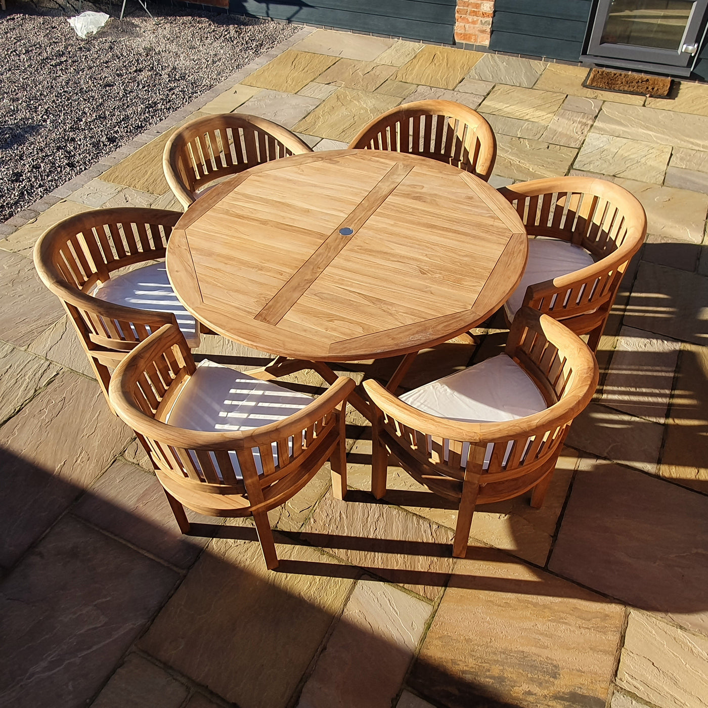 A round Teak 150cm Maximus Table Set with 6 San Francisco Chairs on a paved patio in sunlight.