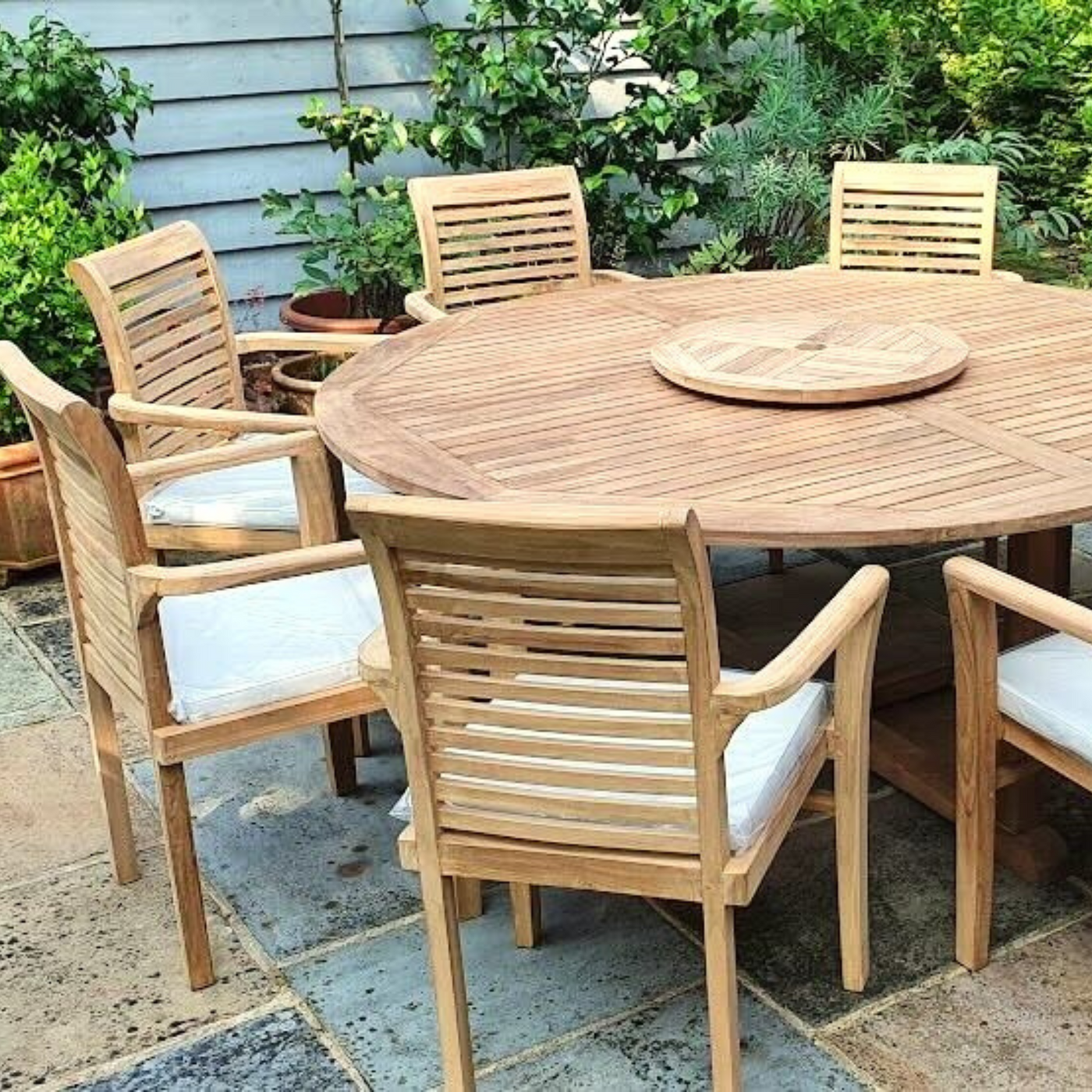 Round Teak 180cm Maximus Round Stacking Set with eight Oxford Stacking Chairs, complete set cushions included, set on a brick floor with lush green plants and a wooden fence in the background.