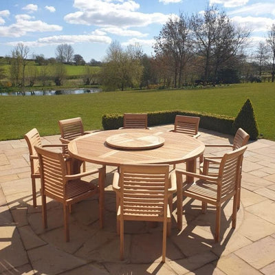 Round Teak 180cm Maximus Round Stacking Set with 8 Oxford Stacking Chairs on a patio, overlooking a sunny landscape with a pond and trees.