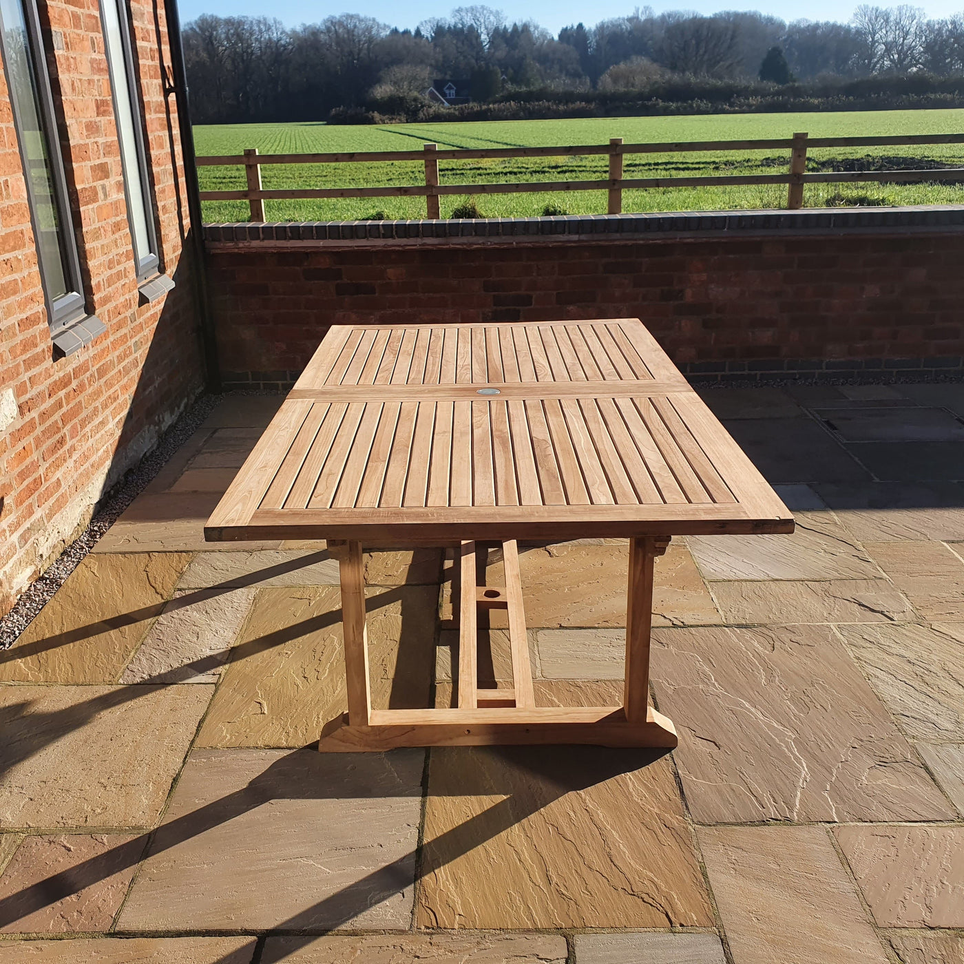 A Teak 2-3m Extending Rectangle Table (10 Seat Oxford Stacking Set) Complete set Cushions included on a stone-paved terrace with a view of a green field and trees under a clear sky.