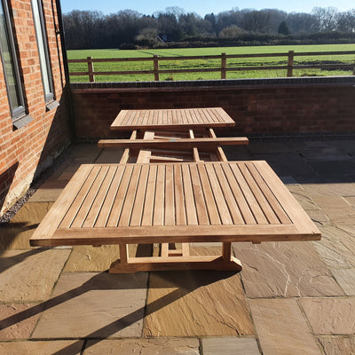 Two Teak 2-3m Extending Rectangle Table (10 Seat Oxford Stacking Set) Complete sets with cushions included on a stone patio beside a brick building, overlooking a grassy field under a clear sky.
